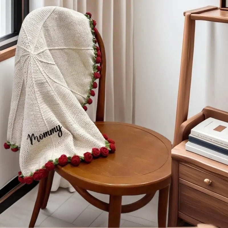 Knitted blanket with 'Mommy' and red flower trim draped over a wooden chair.