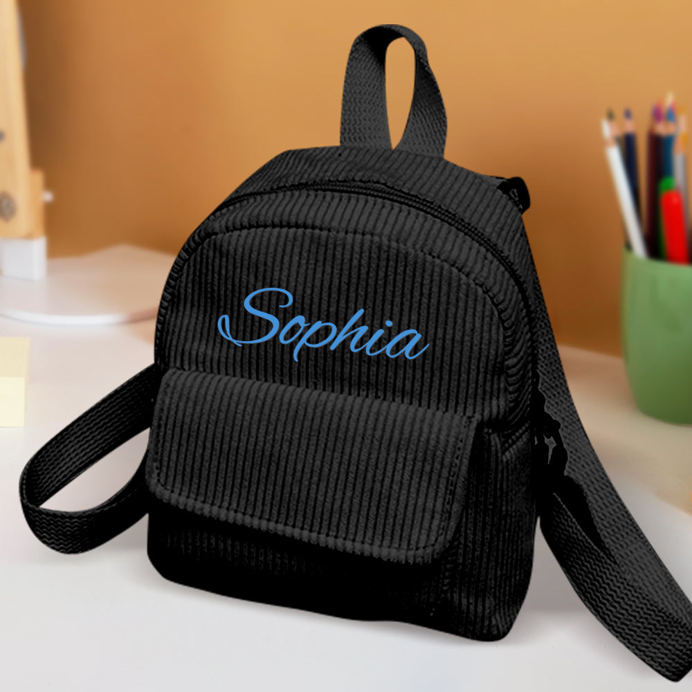 Black mini backpack with 'Sophia' embroidered on it, placed on a surface with a blurred background.