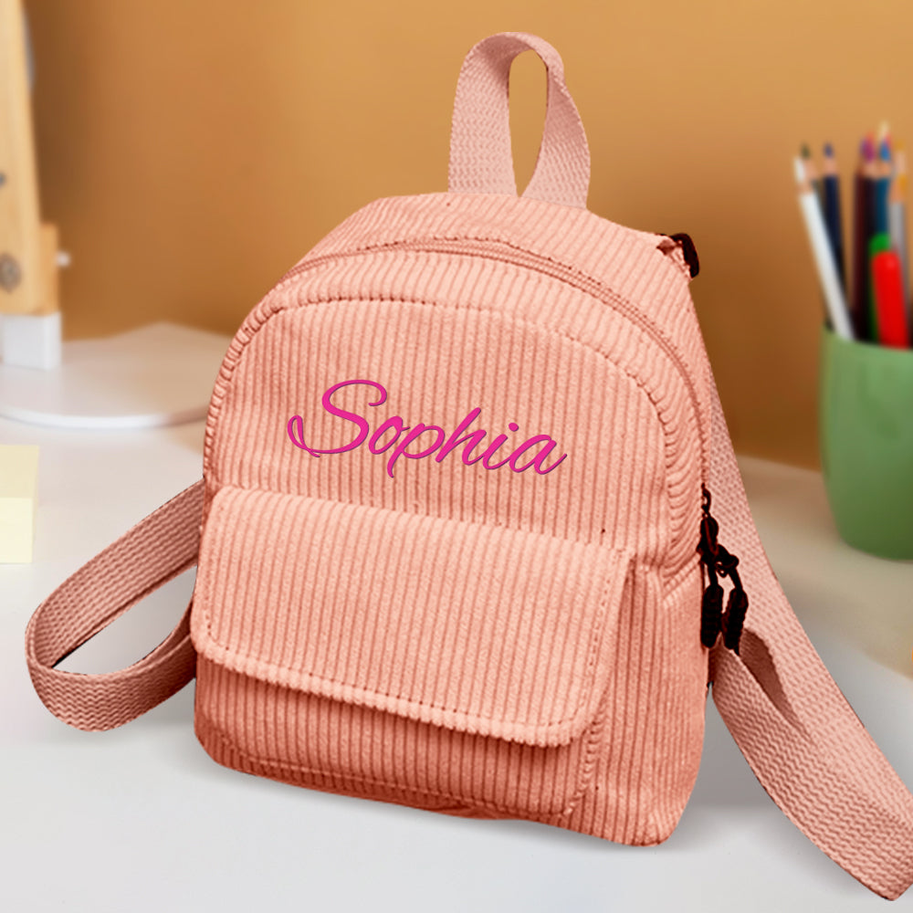 Pink corduroy backpack with 'Sophia' embroidered on it, placed on a surface with a blurred background.