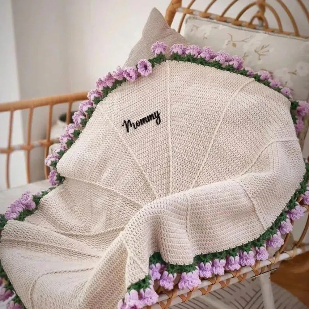 Knitted blanket with 'Mommy' embroidery on a wicker chair with floral decorations.