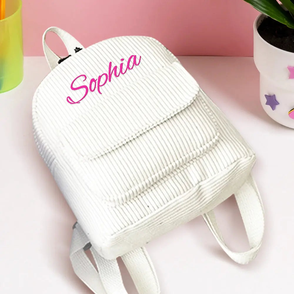 White backpack with 'Sophia' embroidered on it, placed on a pink surface with decorative items.