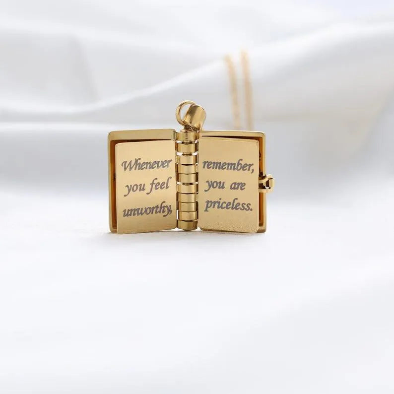 Gold pendant shaped like an open book with engraved text on a white background