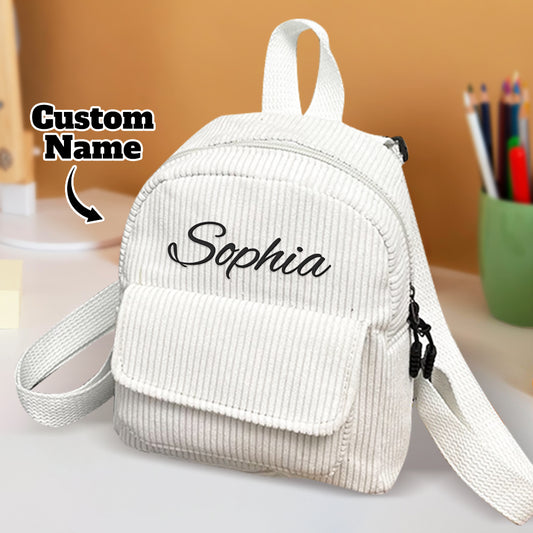 White corduroy backpack with 'Sophia' embroidered on it, placed on a surface with a blurred background.