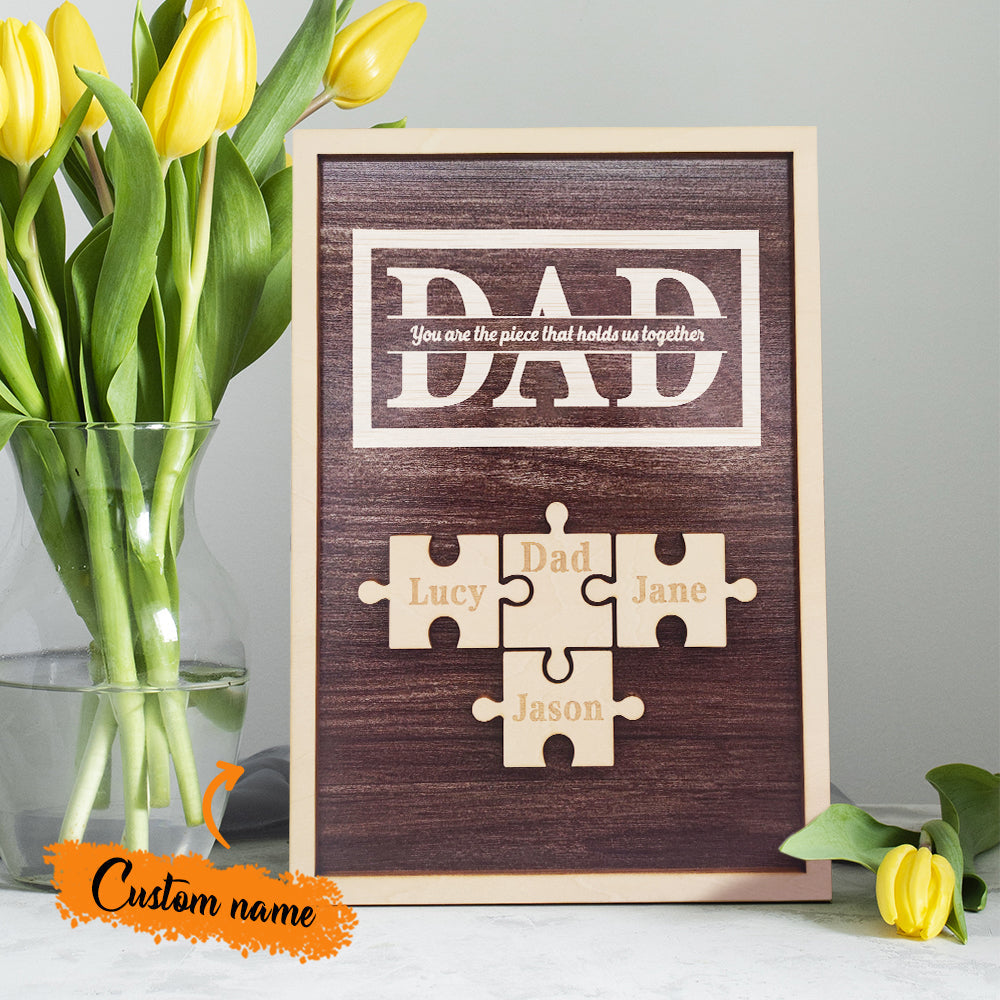 Wooden plaque with puzzle pieces and engraved names, placed next to a vase of yellow tulips.