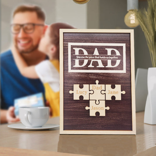 Wooden plaque with puzzle pieces and names on a table with a blurred background of a man and child.