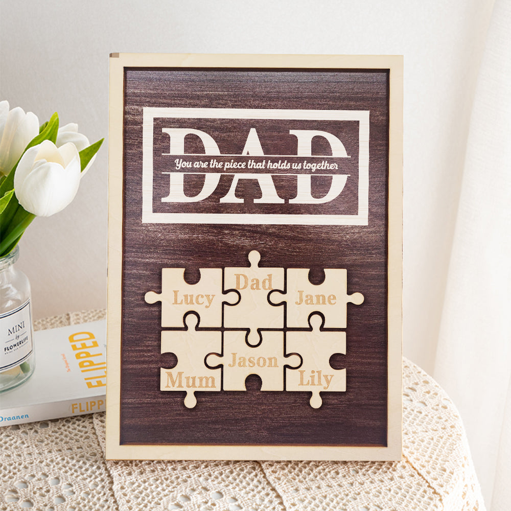Framed puzzle piece art with names on a wooden background, featuring the word 'DAD' and other names.