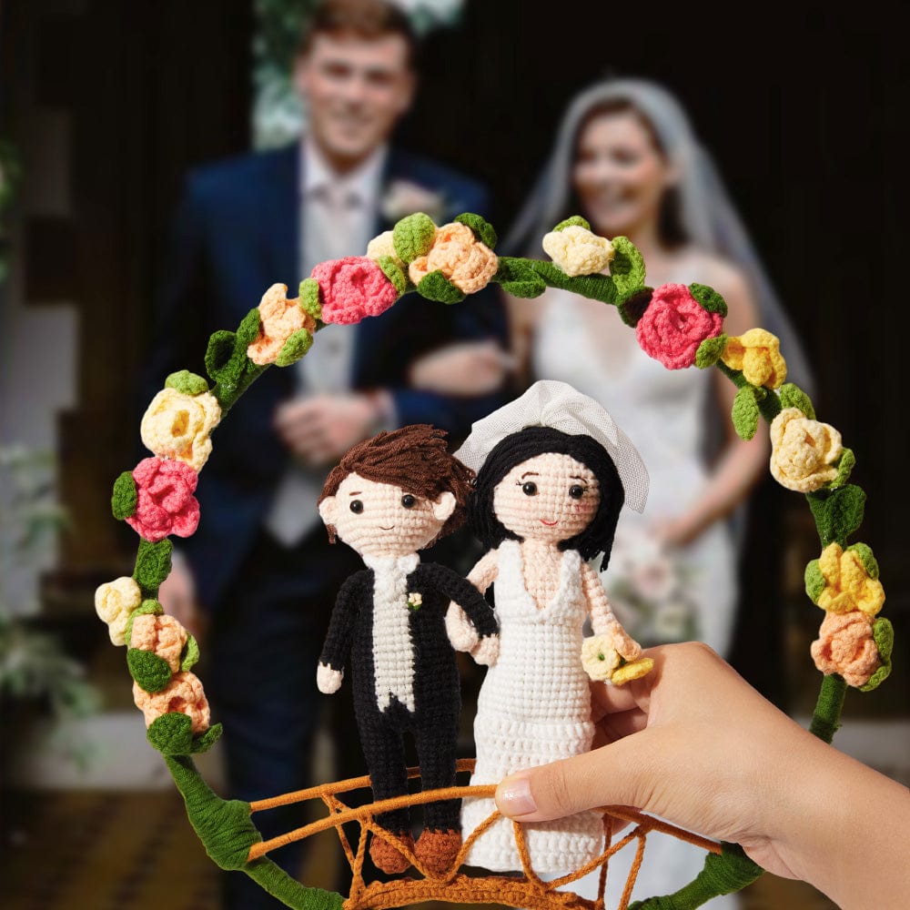 Handmade bride and groom figurines in a floral frame with a blurred wedding couple in the background.
