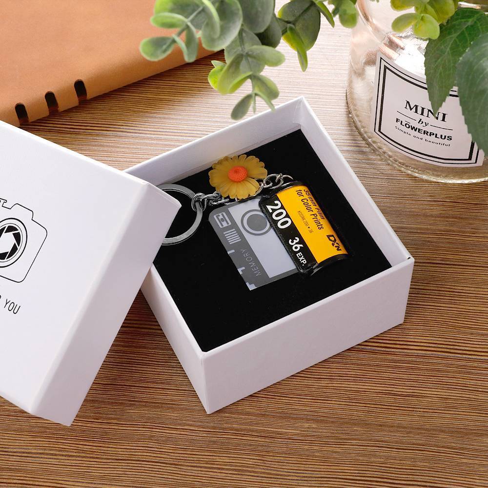 Camera keychain with film roll design in a white box on a wooden surface with plants.