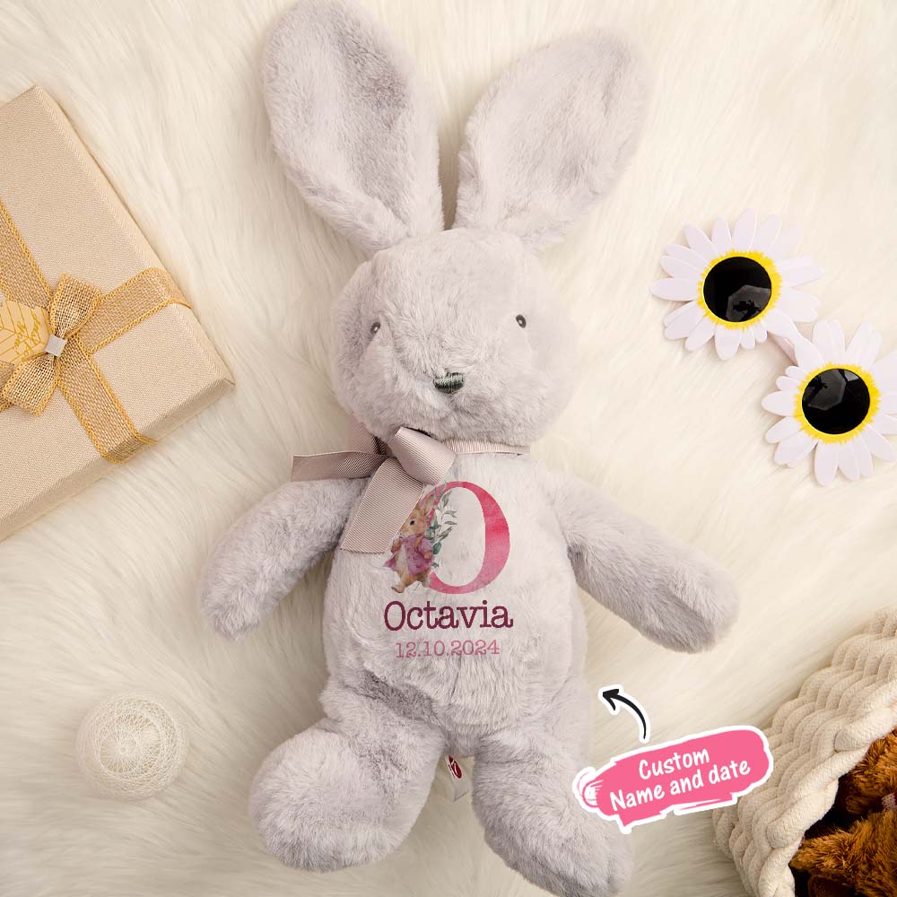Gray plush bunny with 'Octavia' and a date on a soft surface with flowers and a gift.