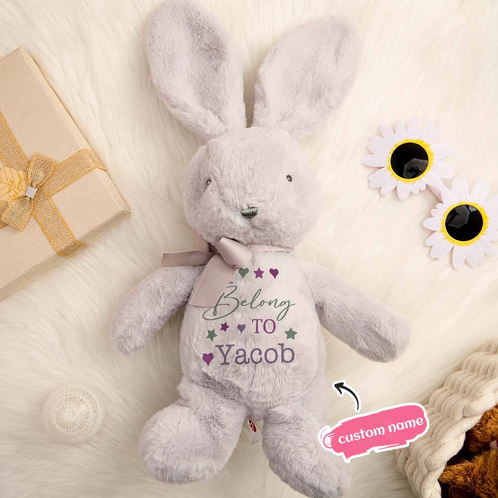 Gray plush bunny with 'Belong To Yacob' text on a soft surface with flowers and gift box.