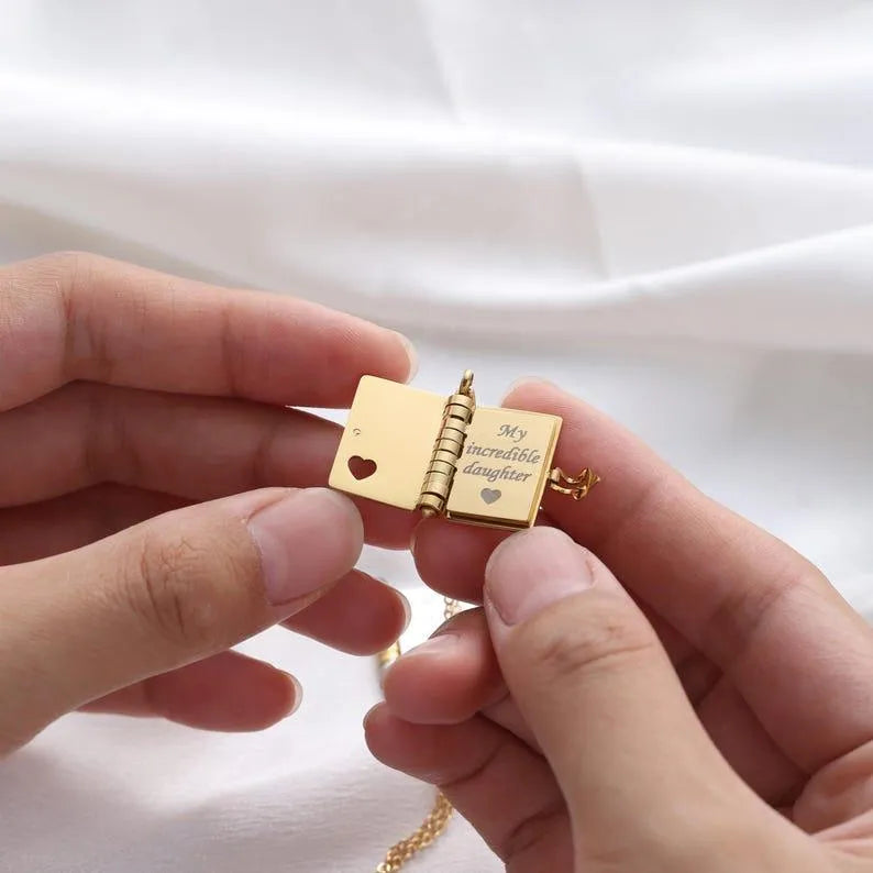 Gold book-shaped pendant with engraved text held between two hands against a white background