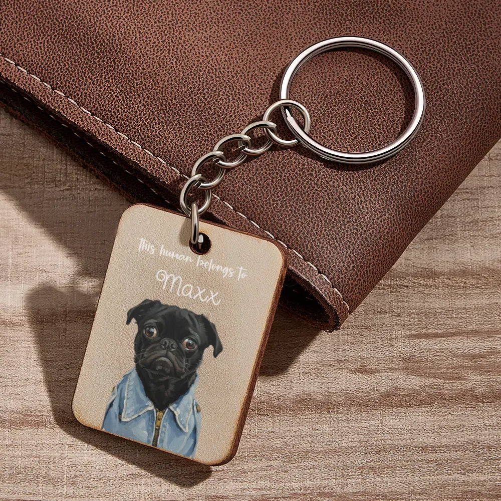 Keychain with a dog illustration and text 'This house belongs to Maxx' on a wooden surface.