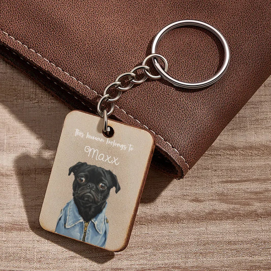 Keychain with a dog illustration and text 'This house belongs to Maxx' on a wooden surface.