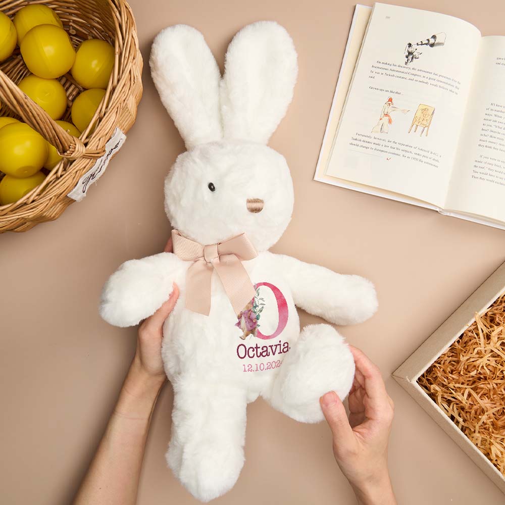 White plush bunny with a pink bow and 'Octavia' on a beige surface with a basket of lemons and an open book.