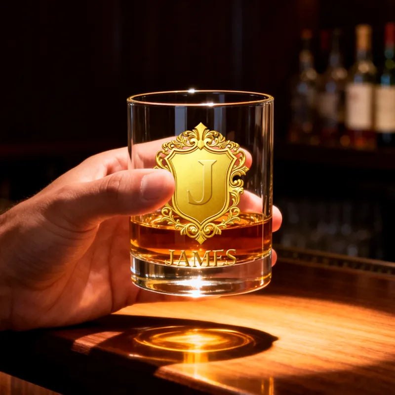 Hand holding a glass with a gold emblem and 'JAMES' engraving, against a bar background.